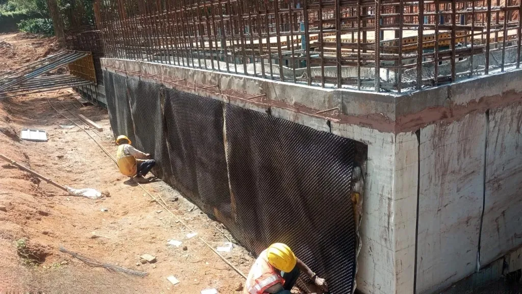 Workers installing studded waterproofing membrane on a basement retaining wall